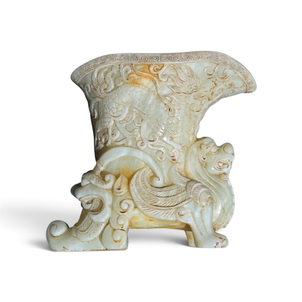 A carved pale celadon nephrite jade rhyton cup featuring relief dragon and mythical beast motifs with subtle russet inclusions.