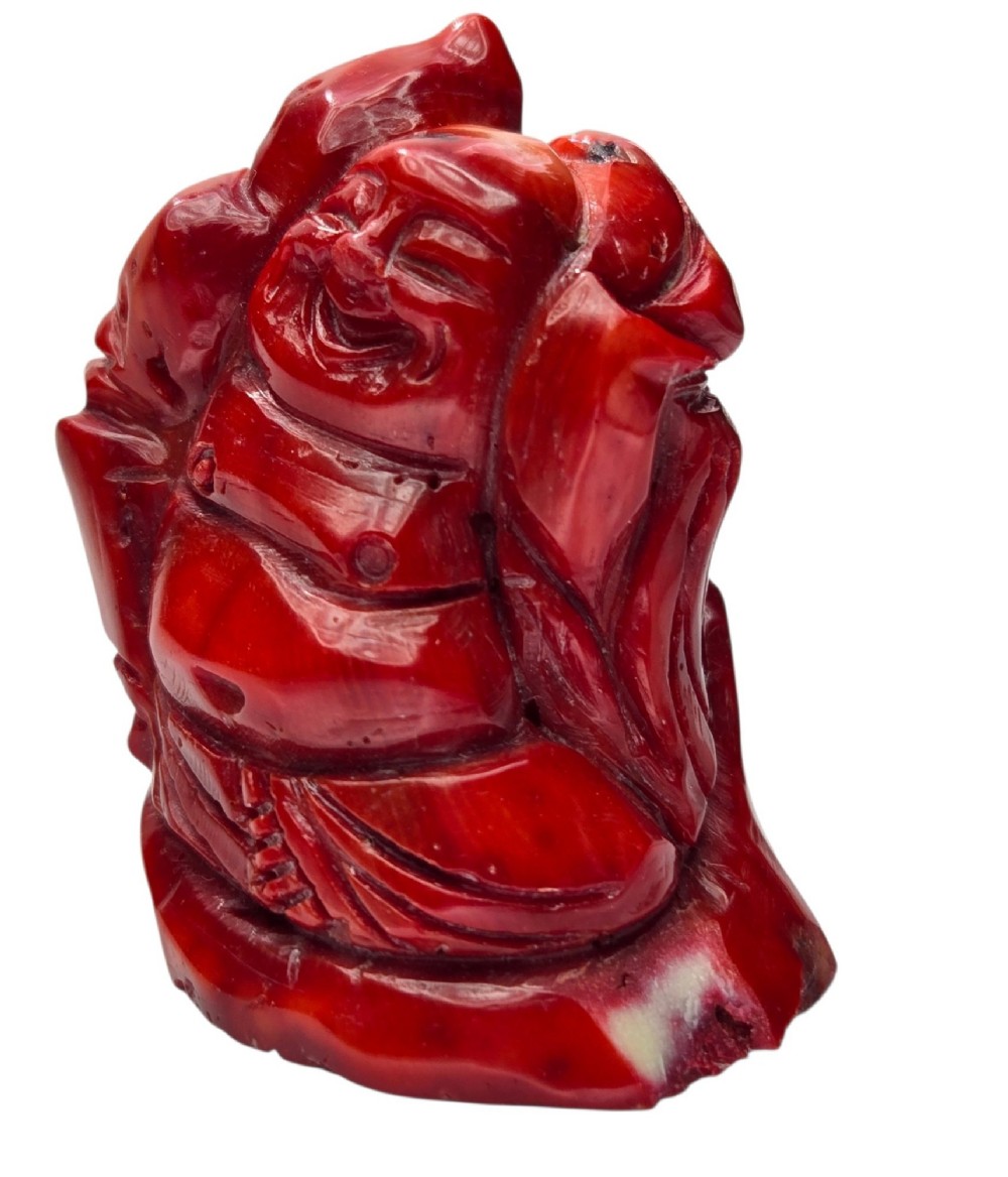 A hand-carved deep red coral sculpture depicting the laughing figure of Budai following the natural shape of a coral branch. View VI