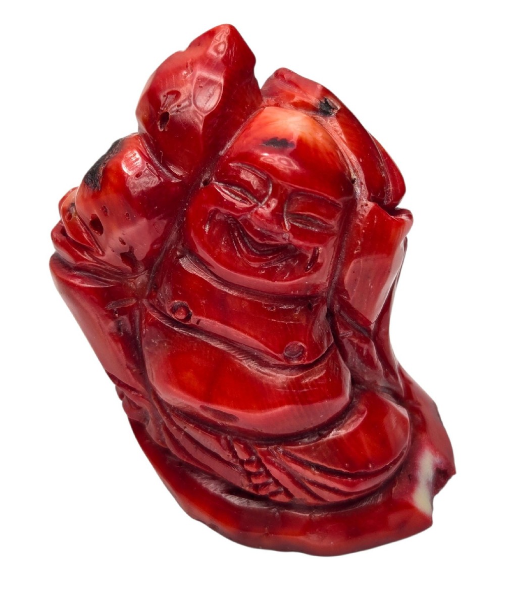 A hand-carved deep red coral sculpture depicting the laughing figure of Budai following the natural shape of a coral branch. View IX