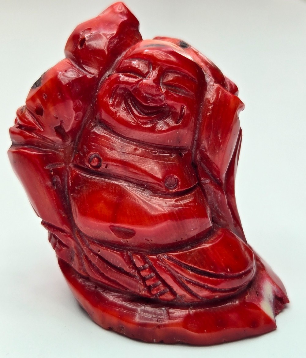 A hand-carved deep red coral sculpture depicting the laughing figure of Budai following the natural shape of a coral branch. View XV