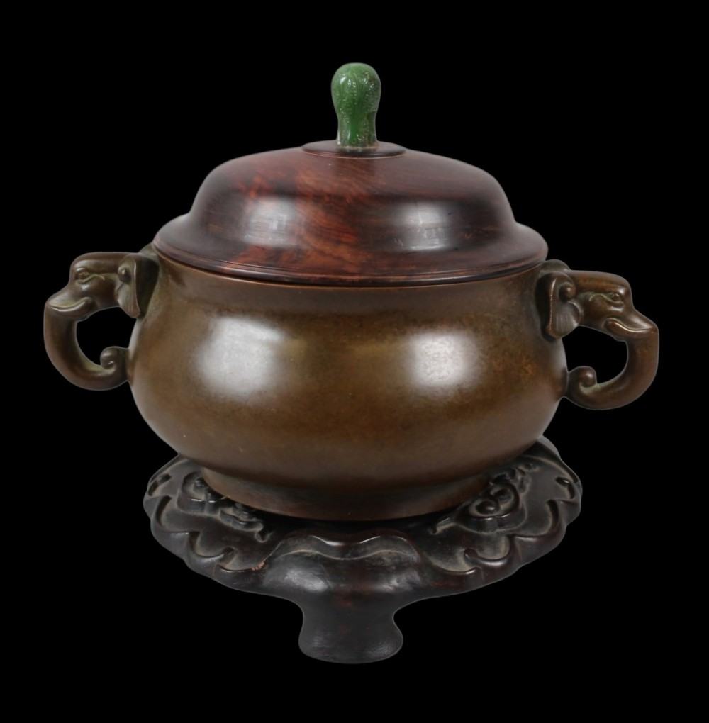 A rounded dark bronze Chinese censer with elephant head handles, topped with a polished wood lid featuring a green jade finial, resting on a carved hardwood stand. View II