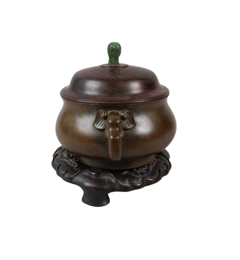 A rounded dark bronze Chinese censer with elephant head handles, topped with a polished wood lid featuring a green jade finial, resting on a carved hardwood stand. View III