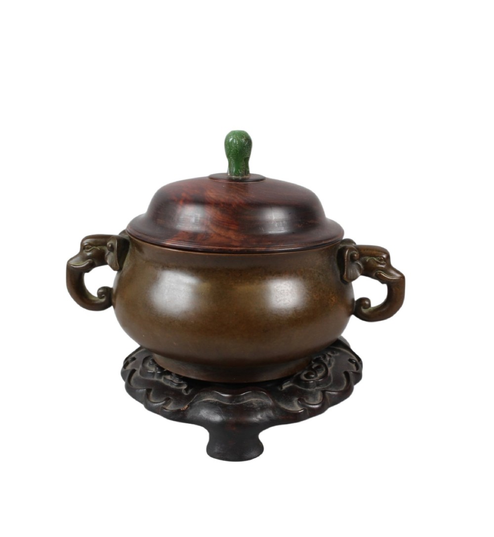 A rounded dark bronze Chinese censer with elephant head handles, topped with a polished wood lid featuring a green jade finial, resting on a carved hardwood stand. View IV