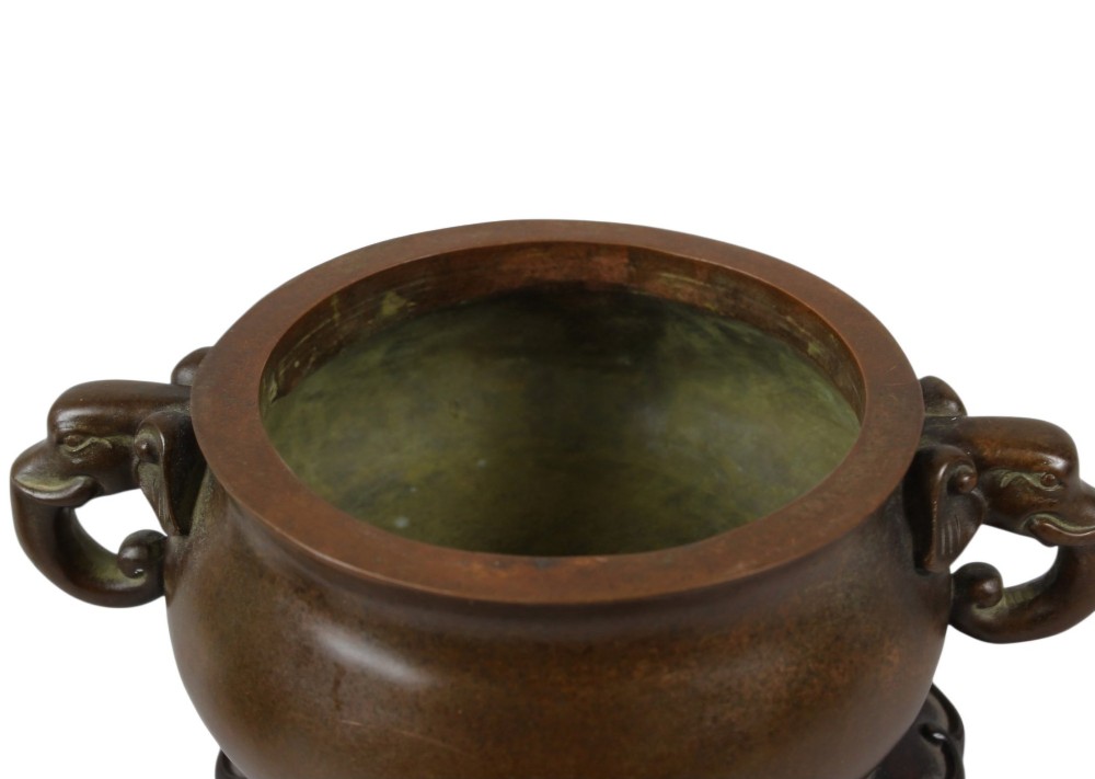 A rounded dark bronze Chinese censer with elephant head handles, topped with a polished wood lid featuring a green jade finial, resting on a carved hardwood stand. View VI