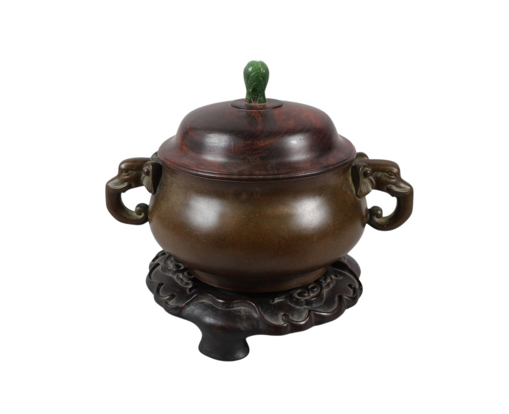 A rounded dark bronze Chinese censer with elephant head handles, topped with a polished wood lid featuring a green jade finial, resting on a carved hardwood stand. View IX