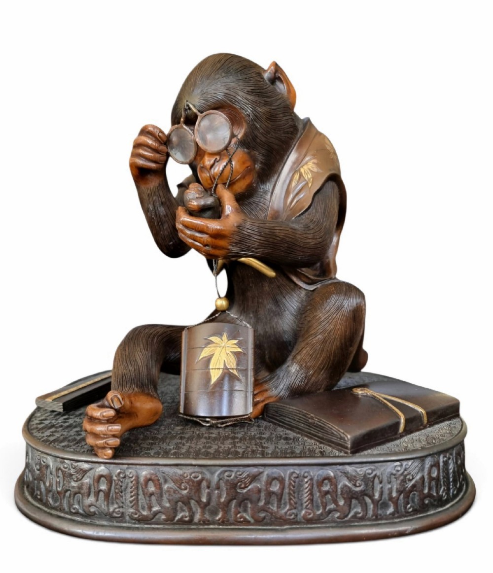 A dark bronze Japanese okimono sculpture depicting an anthropomorphic monkey wearing spectacles, seated with a book and inro.