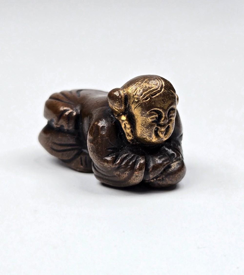 A dark bronze cast sculpture of a reclining child with gilded details on the head and face, resting on a flat base. View X