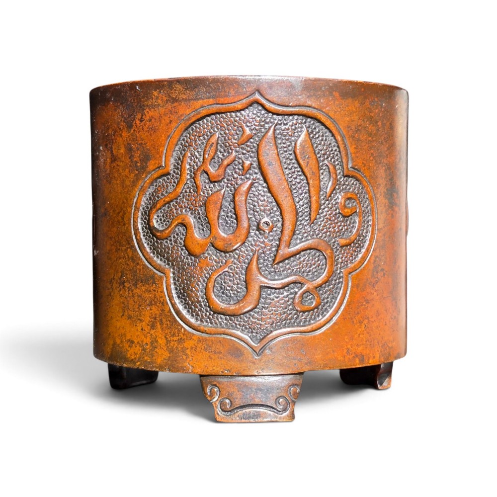 A cylindrical warm brown bronze tripod censer featuring a raised lobed cartouche with Arabic calligraphy set against a textured ground.
