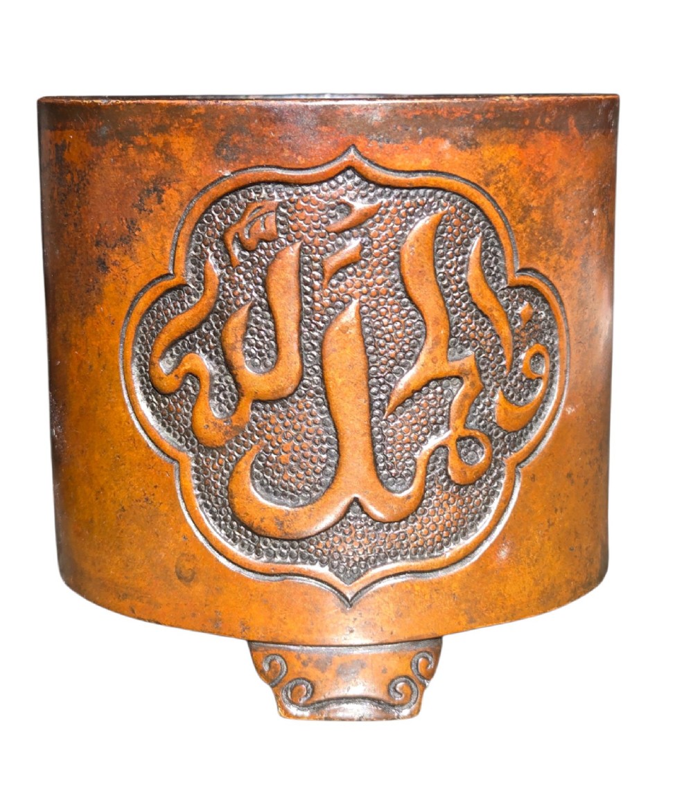 A cylindrical warm brown bronze tripod censer featuring a raised lobed cartouche with Arabic calligraphy set against a textured ground. View V