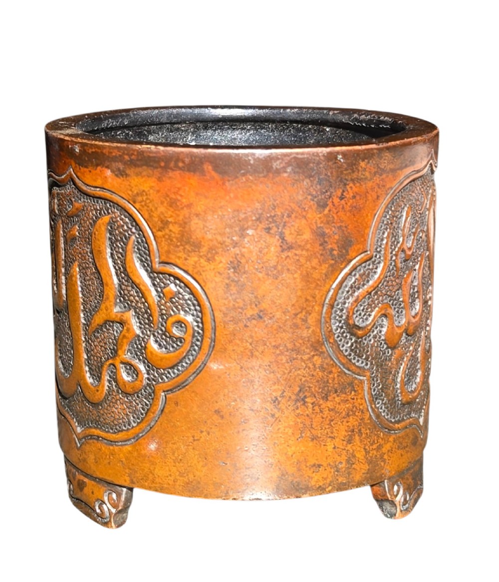 A cylindrical warm brown bronze tripod censer featuring a raised lobed cartouche with Arabic calligraphy set against a textured ground. View IX