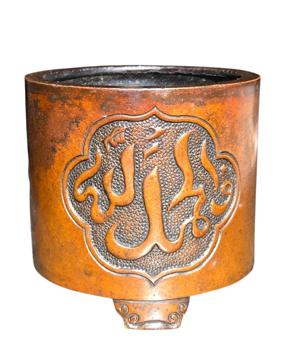 A cylindrical warm brown bronze tripod censer featuring a raised lobed cartouche with Arabic calligraphy set against a textured ground. View X