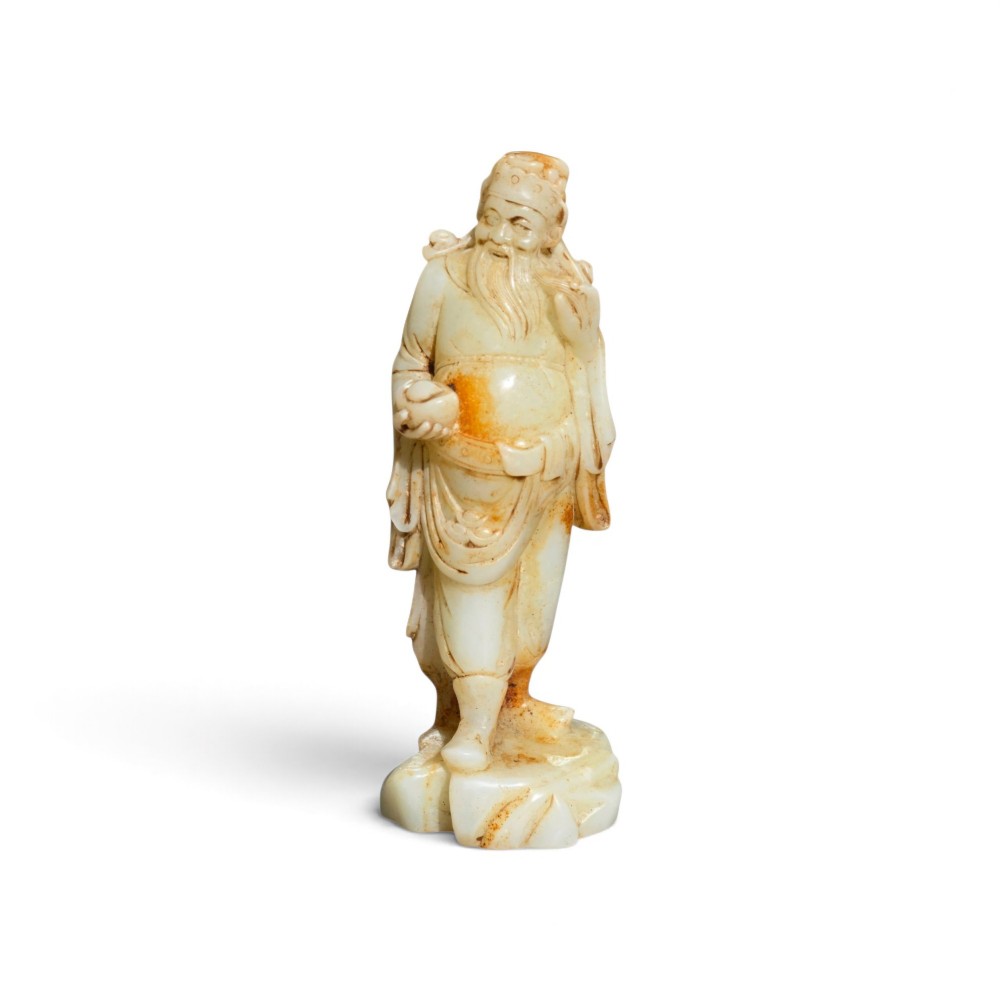 A carved pale celadon and russet nephrite jade standing figure of an immortal holding a vessel on a carved rockwork base. View II