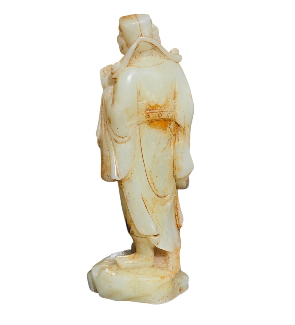 A carved pale celadon and russet nephrite jade standing figure of an immortal holding a vessel on a carved rockwork base. View VI