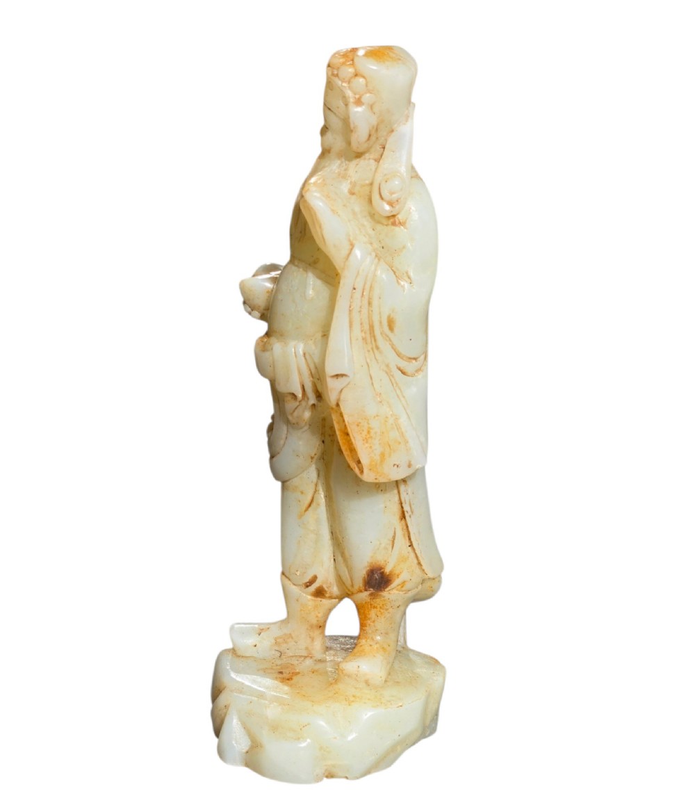 A carved pale celadon and russet nephrite jade standing figure of an immortal holding a vessel on a carved rockwork base. View VII