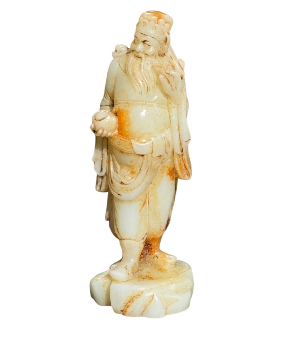 A carved pale celadon and russet nephrite jade standing figure of an immortal holding a vessel on a carved rockwork base. View IX