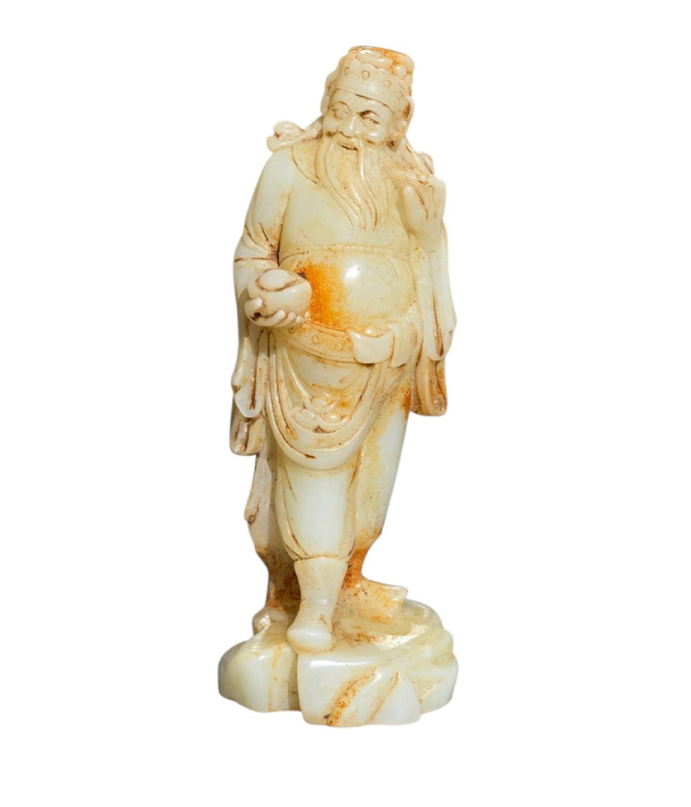 A carved pale celadon and russet nephrite jade standing figure of an immortal holding a vessel on a carved rockwork base. View X