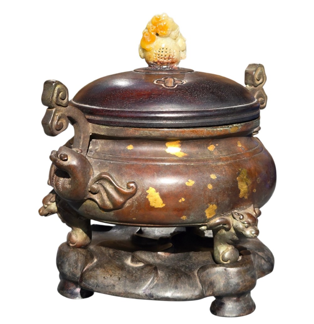 An 18th-century Chinese gilt-splashed bronze tripod censer featuring scroll handles, animal-mask legs, a dark hardwood cover topped with a carved jade finial, and a fitted bronze stand. View X