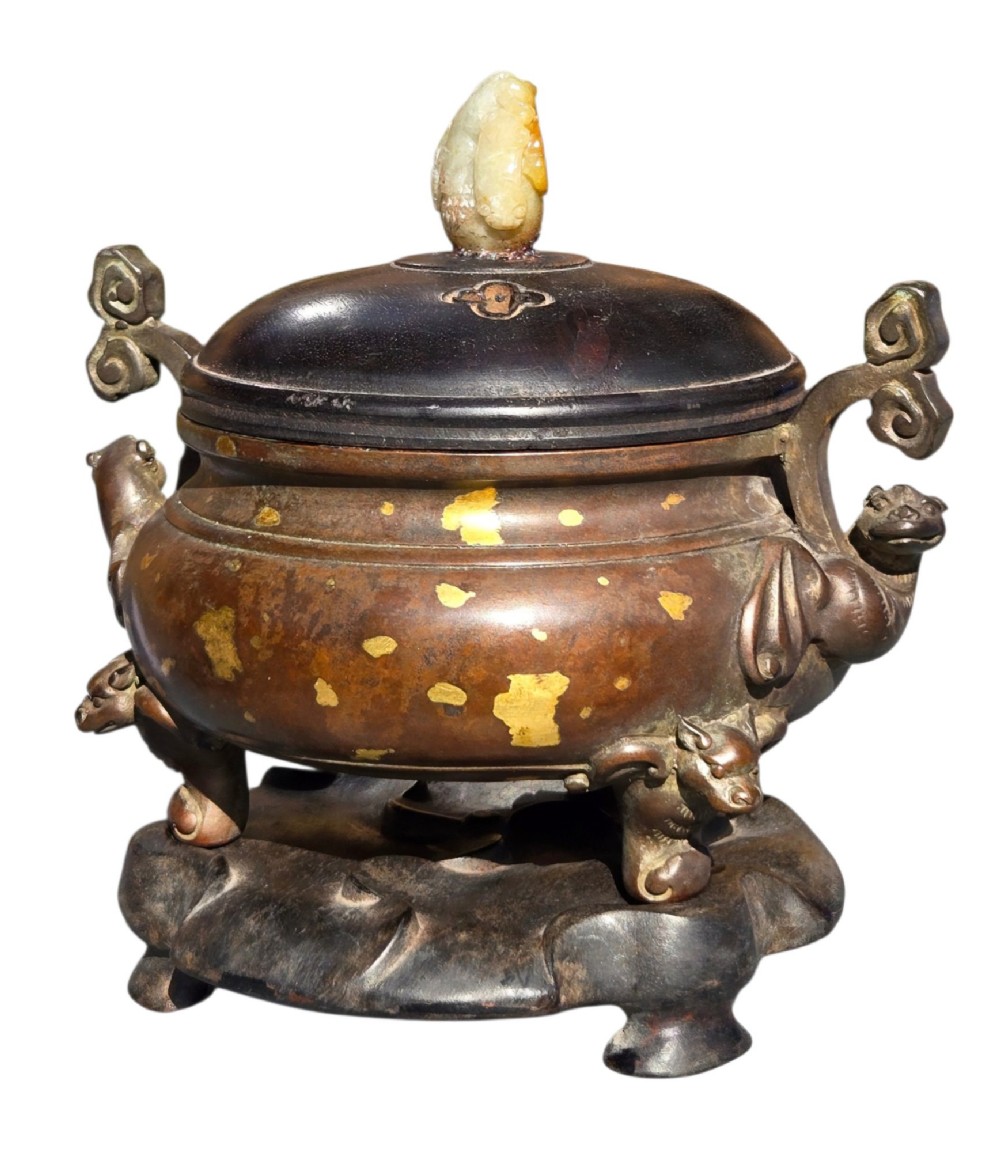 An 18th-century Chinese gilt-splashed bronze tripod censer featuring scroll handles, animal-mask legs, a dark hardwood cover topped with a carved jade finial, and a fitted bronze stand. View XIV