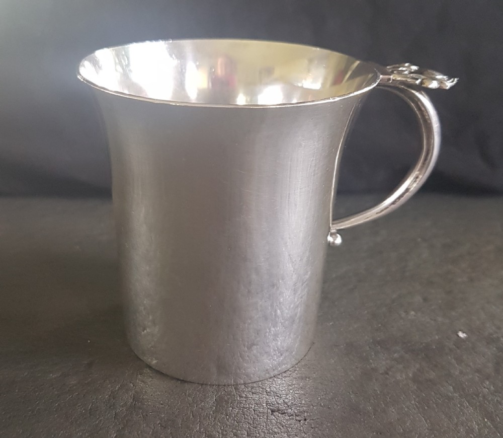 A sterling silver christening mug featuring a slightly flared rim, planished surface texture, and a curved loop handle detailed with an acorn motif.