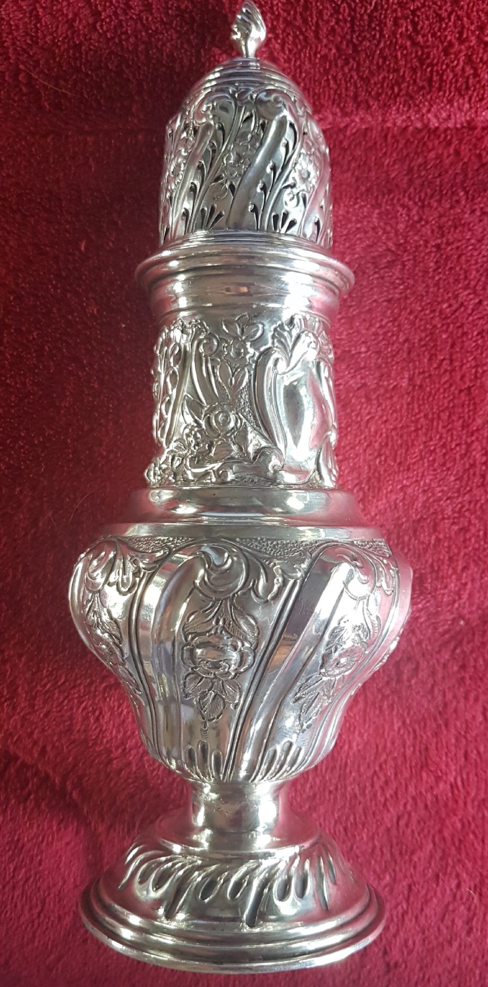 An ornate sterling silver sugar caster featuring intricate repousse floral motifs, a pierced decorative dome cover with a flame finial, and a fluted circular base.