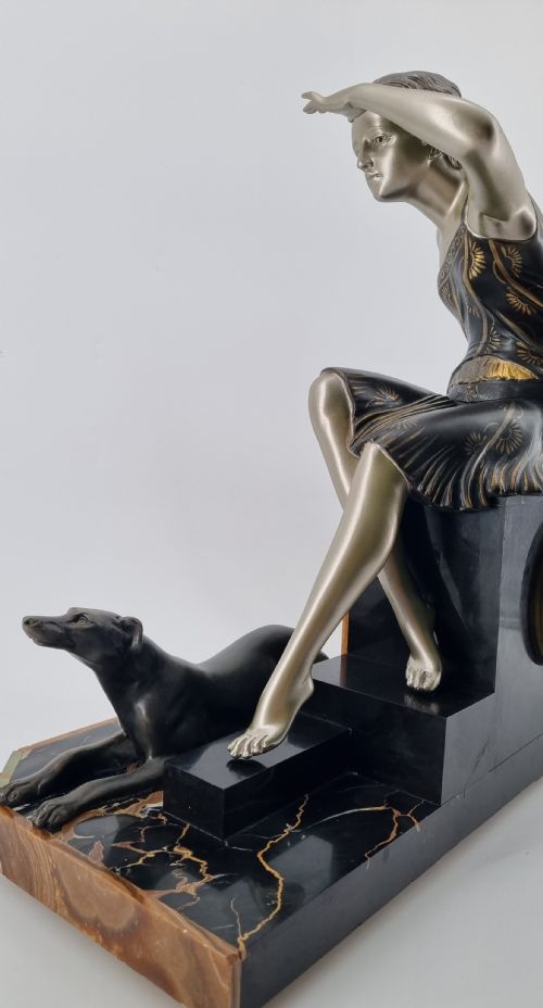 fig 1. An Art Deco mantel clock featuring a silvered metal figure of a seated woman flanked by two greyhounds atop a multi-coloured green and brown marble base with an octagonal dial.