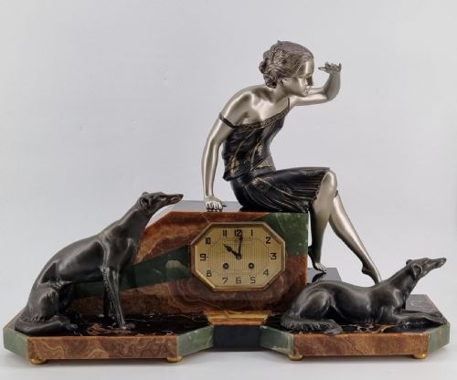 fig 2. An Art Deco mantel clock featuring a silvered metal figure of a seated woman flanked by two greyhounds atop a multi-coloured green and brown marble base with an octagonal dial.