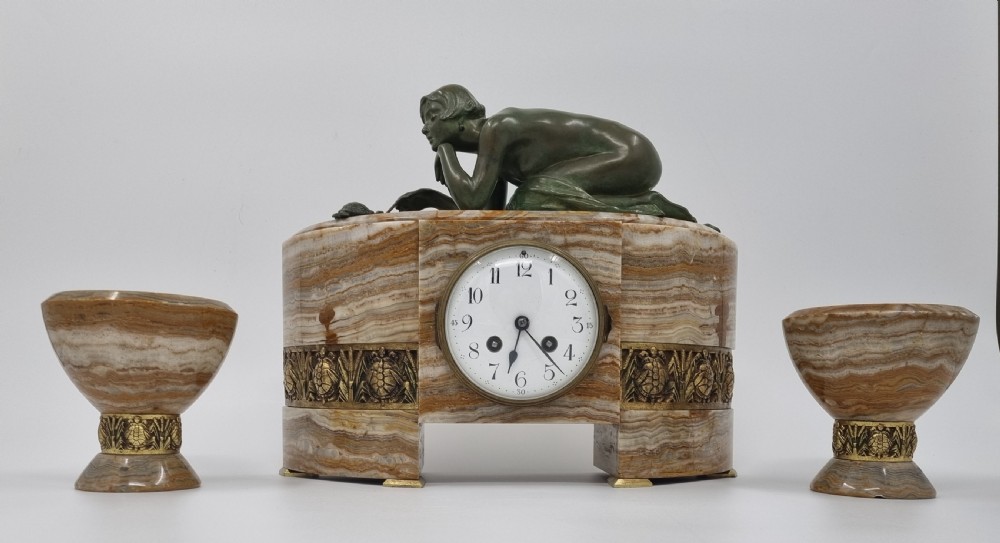 An Art Deco clock garniture set featuring a carved onyx mantel clock topped with a green patinated bronze sculpture of a kneeling woman, accompanied by two matching onyx cups. View II