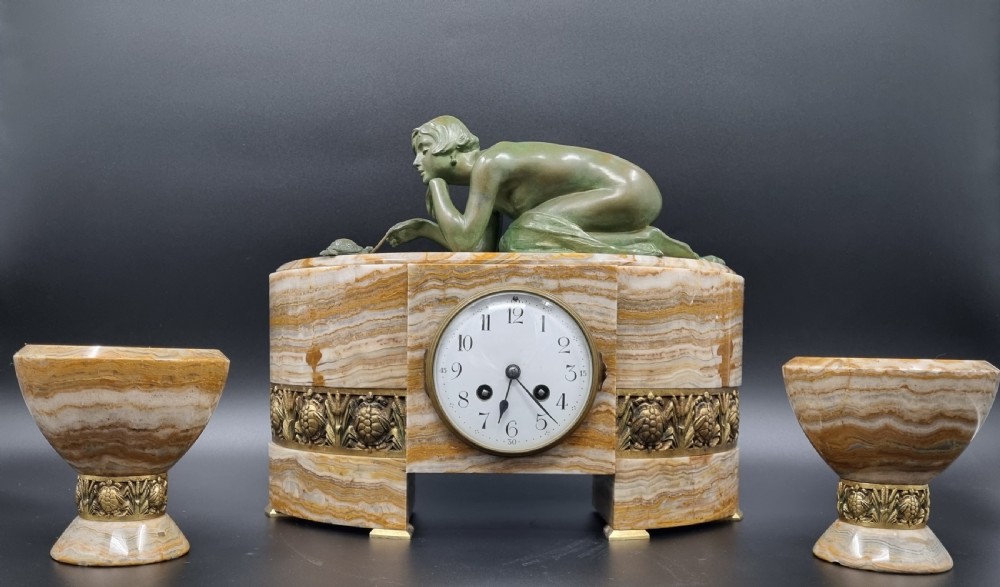 An Art Deco clock garniture set featuring a carved onyx mantel clock topped with a green patinated bronze sculpture of a kneeling woman, accompanied by two matching onyx cups. View XVI