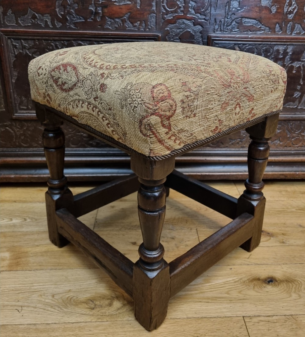 A square 17th-century oak stool featuring turned legs, rectangular box stretchers, and a patterned brown fabric upholstered seat. View V