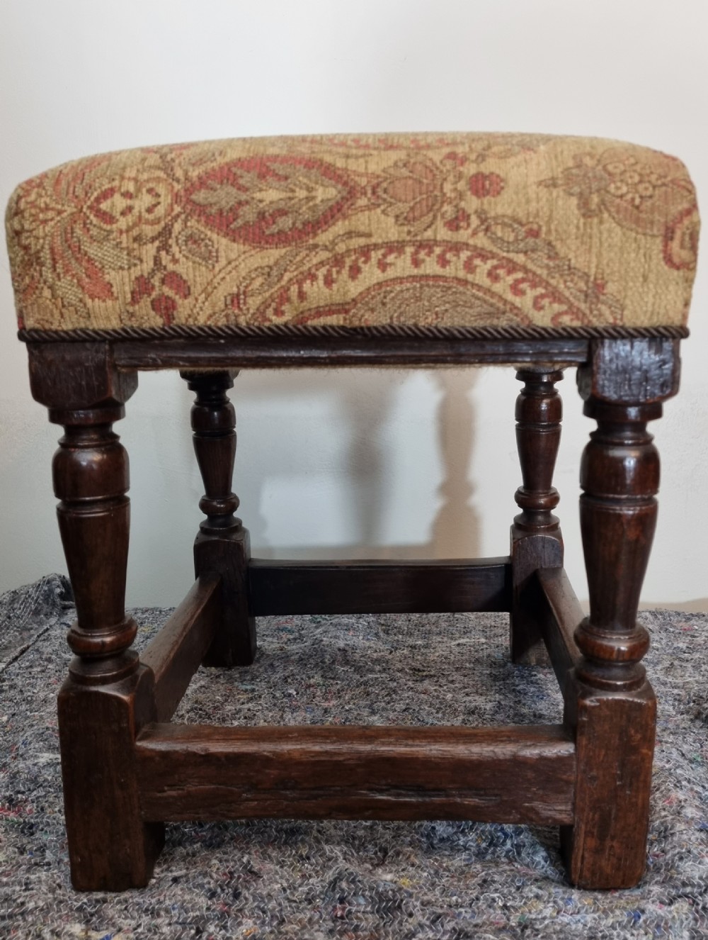 A square 17th-century oak stool featuring turned legs, rectangular box stretchers, and a patterned brown fabric upholstered seat. View VI