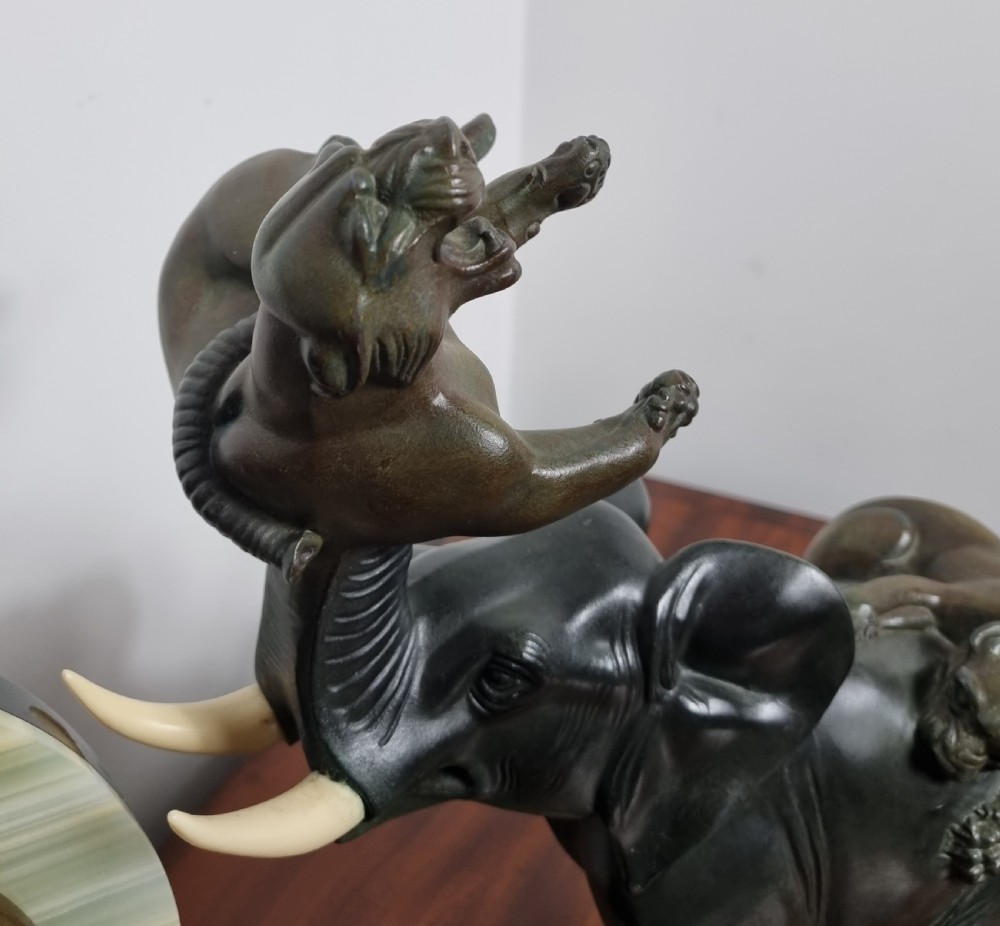 An Art Deco mantel clock featuring a circular green onyx dial flanked by dark patinated spelter sculptures of an elephant and panthers on a layered marble base. View VII