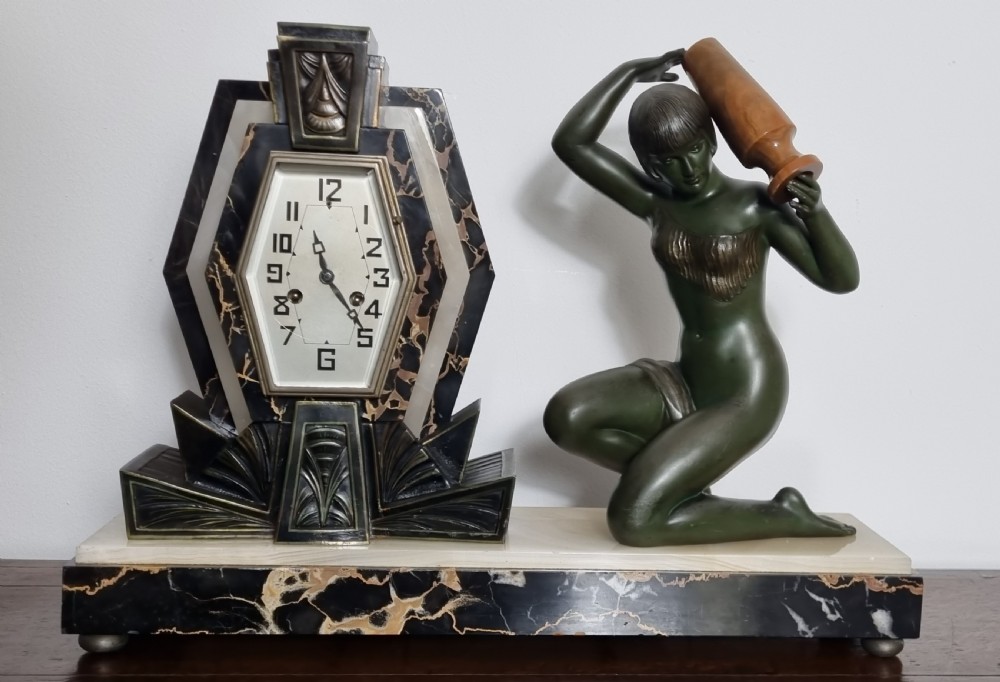An Art Deco illuminated mantel clock featuring a dark figure of a kneeling woman holding a vessel beside a geometric hexagonal clock face mounted on a black and white veined marble base. View VI