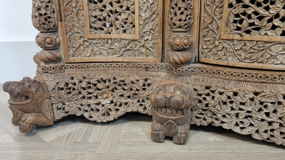 A 19th-century Anglo-Indian intricately carved teak wood cabinet featuring openwork floral lattice panels, figure pilasters, arched cresting, and carved animal feet. View VI