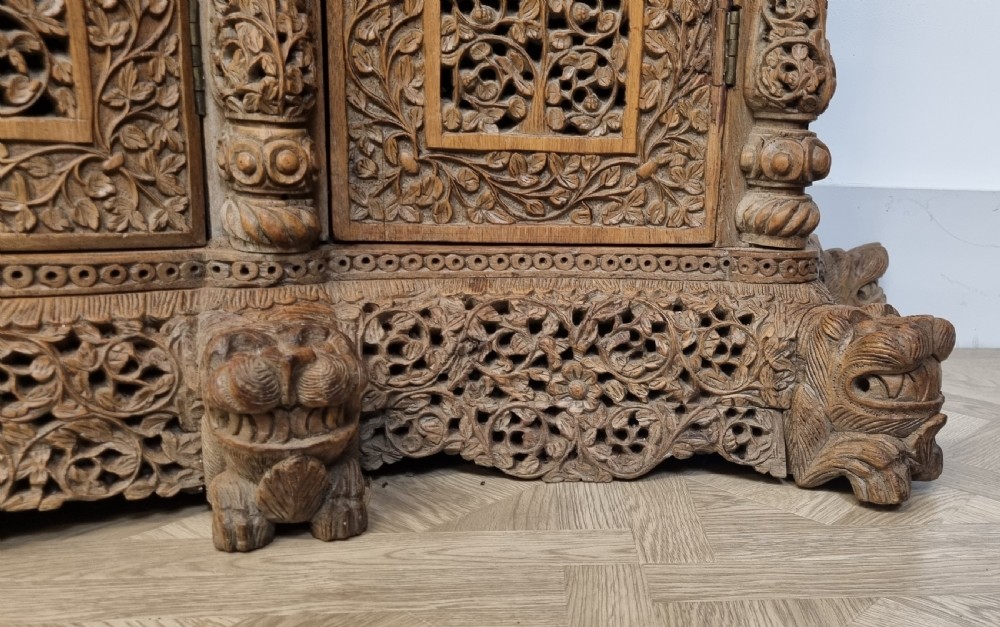 A 19th-century Anglo-Indian intricately carved teak wood cabinet featuring openwork floral lattice panels, figure pilasters, arched cresting, and carved animal feet. View XIV