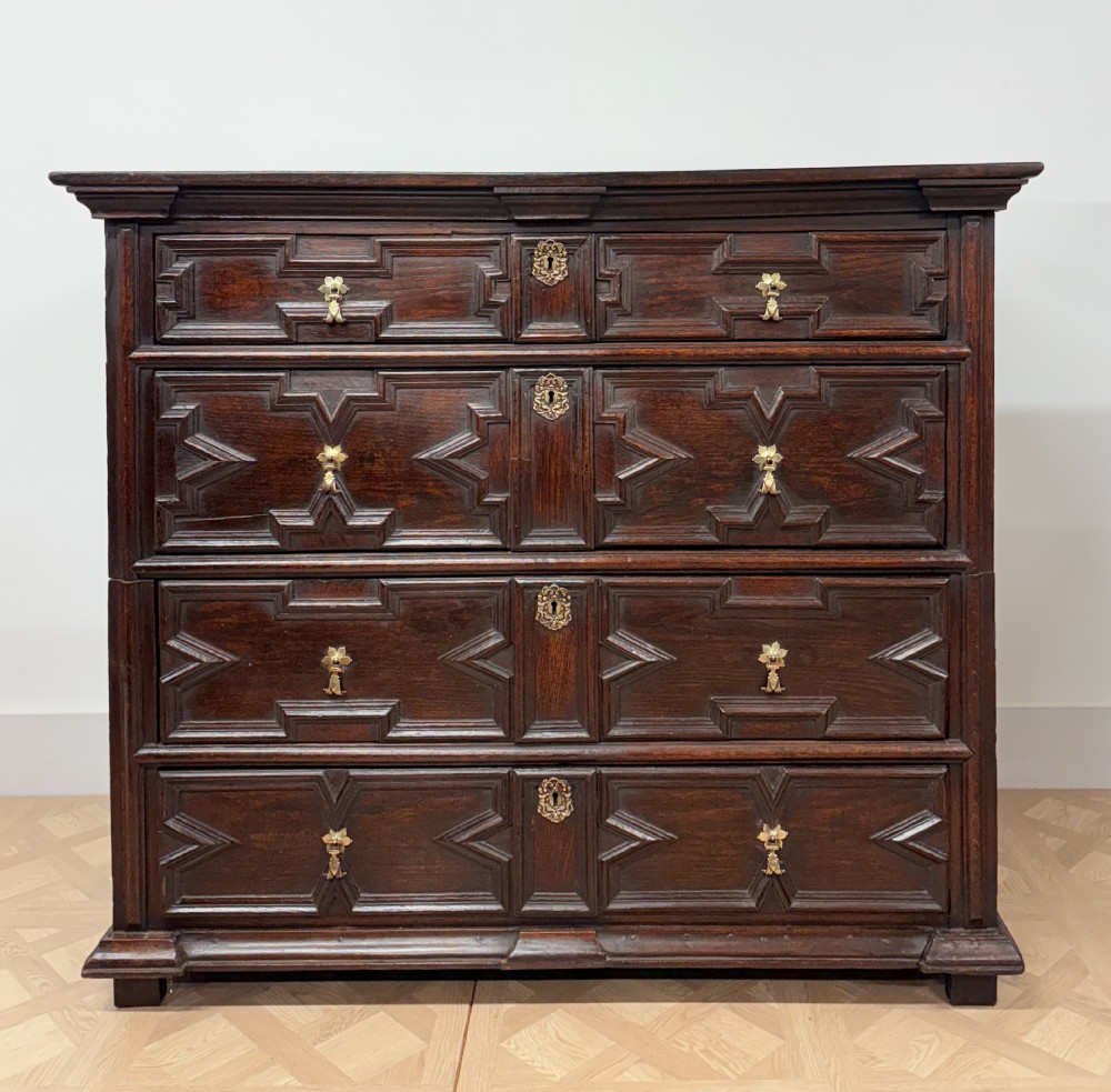 A seventeenth-century dark oak chest of four long drawers featuring geometric panel mouldings and brass teardrop handles.