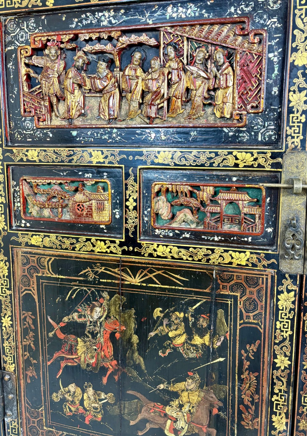 Late 19th-century Chinese black lacquer cabinet featuring intricate hand-carved polychrome figures, gilded panels of warriors on horseback, and brass door fittings. View VII