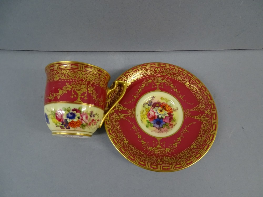 A Royal Worcester demitasse cup and matching saucer featuring crimson ground decoration, gilded floral swags, and a hand-painted floral centre. View II