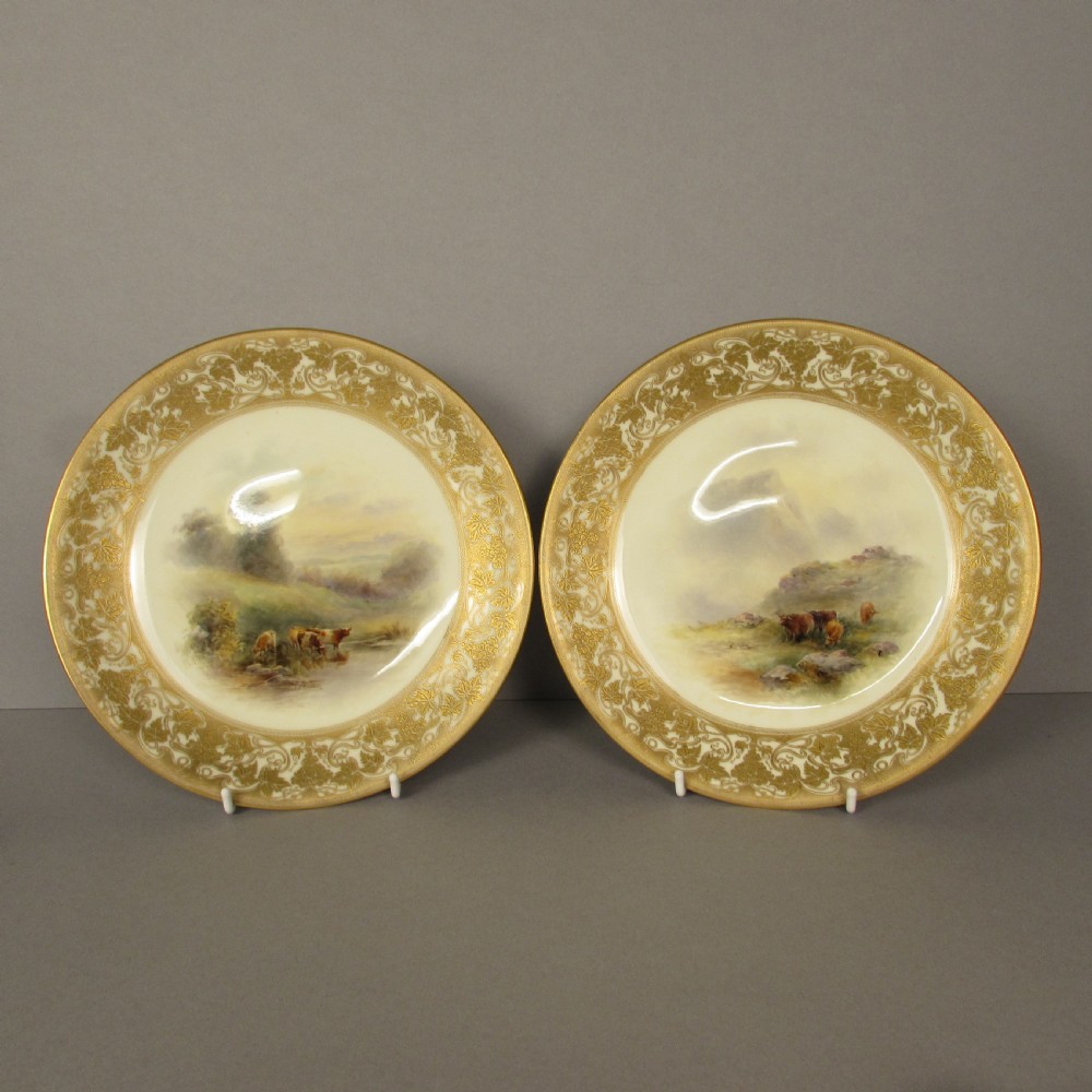 A pair of round Royal Worcester porcelain cabinet plates featuring hand-painted scenes of cattle in landscapes, bordered by ornate gold scrollwork gilding on a cream ground.