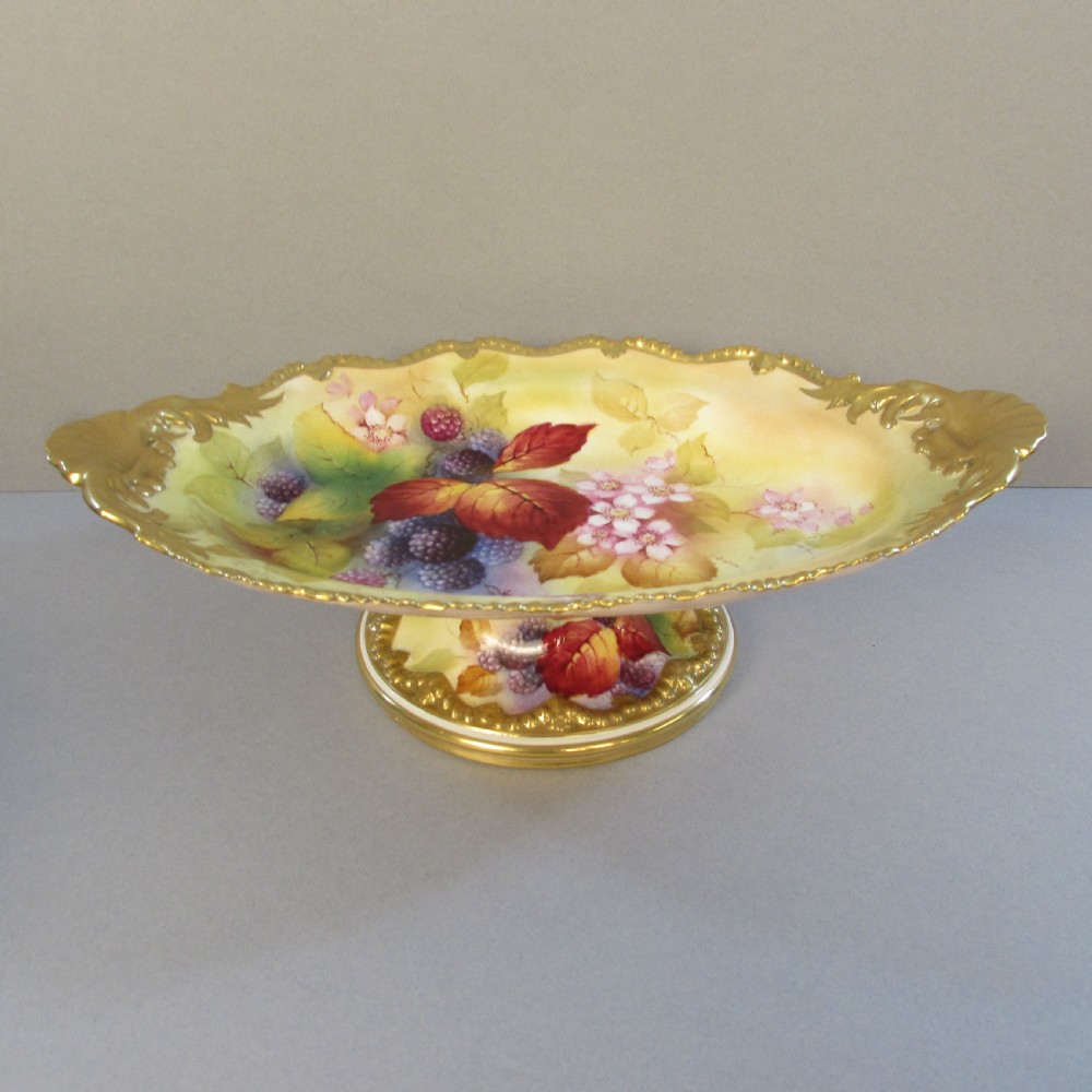 An oval hand-painted porcelain comport featuring a motif of blackberries and autumn leaves with gilded handles and a pedestal base.