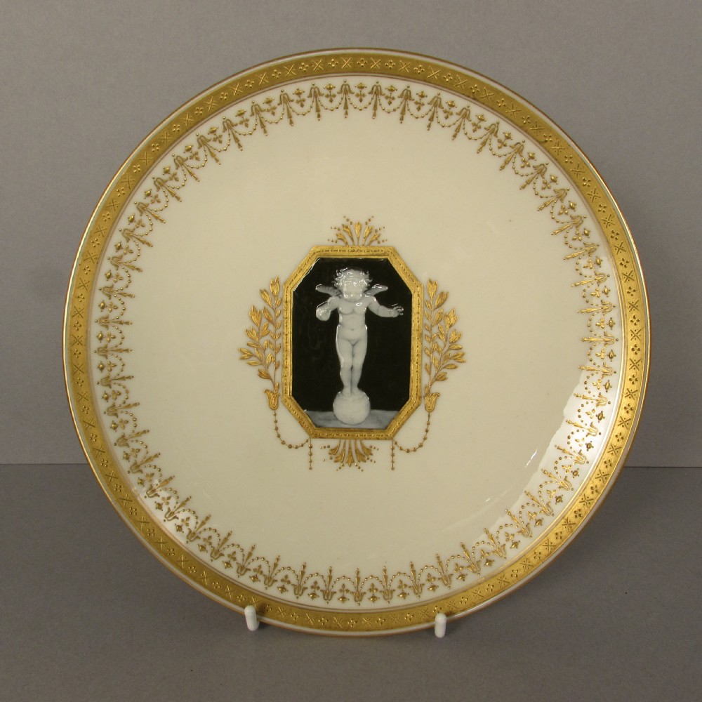 A circular cream porcelain cabinet plate featuring a central black octagonal medallion with a white relief cherub figure framed by gold leaf swags and an ornate gilded border.