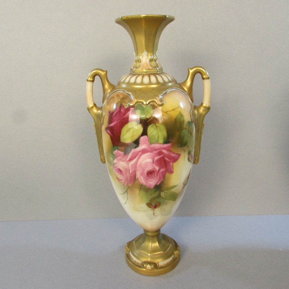 A hand-painted Royal Worcester porcelain vase featuring pink roses, twin gilded handles, and a flared neck on a pedestal base.