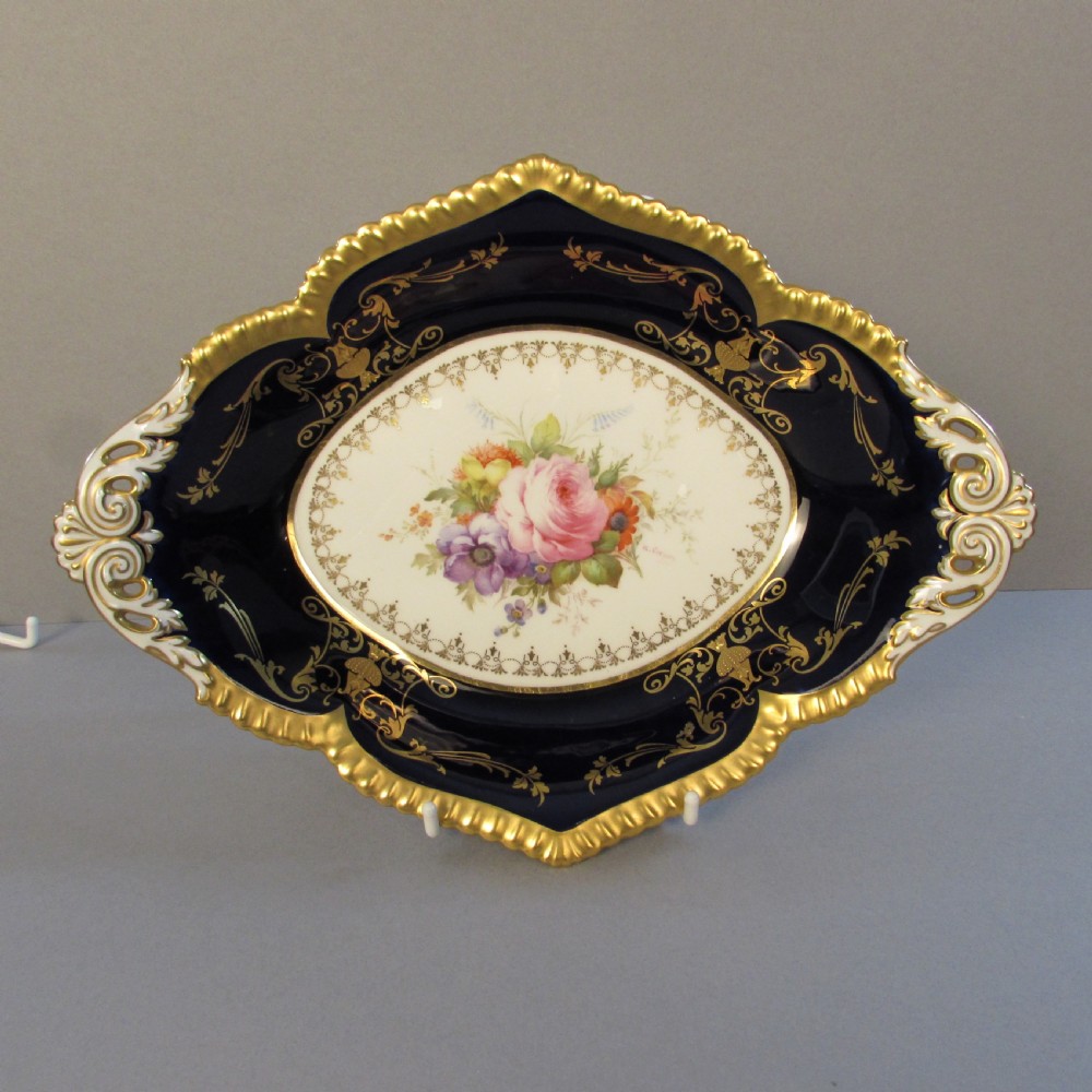 An oval porcelain dish featuring a cobalt blue border, gold scroll decoration, pierced handles, and a central hand-painted floral bouquet reserve.