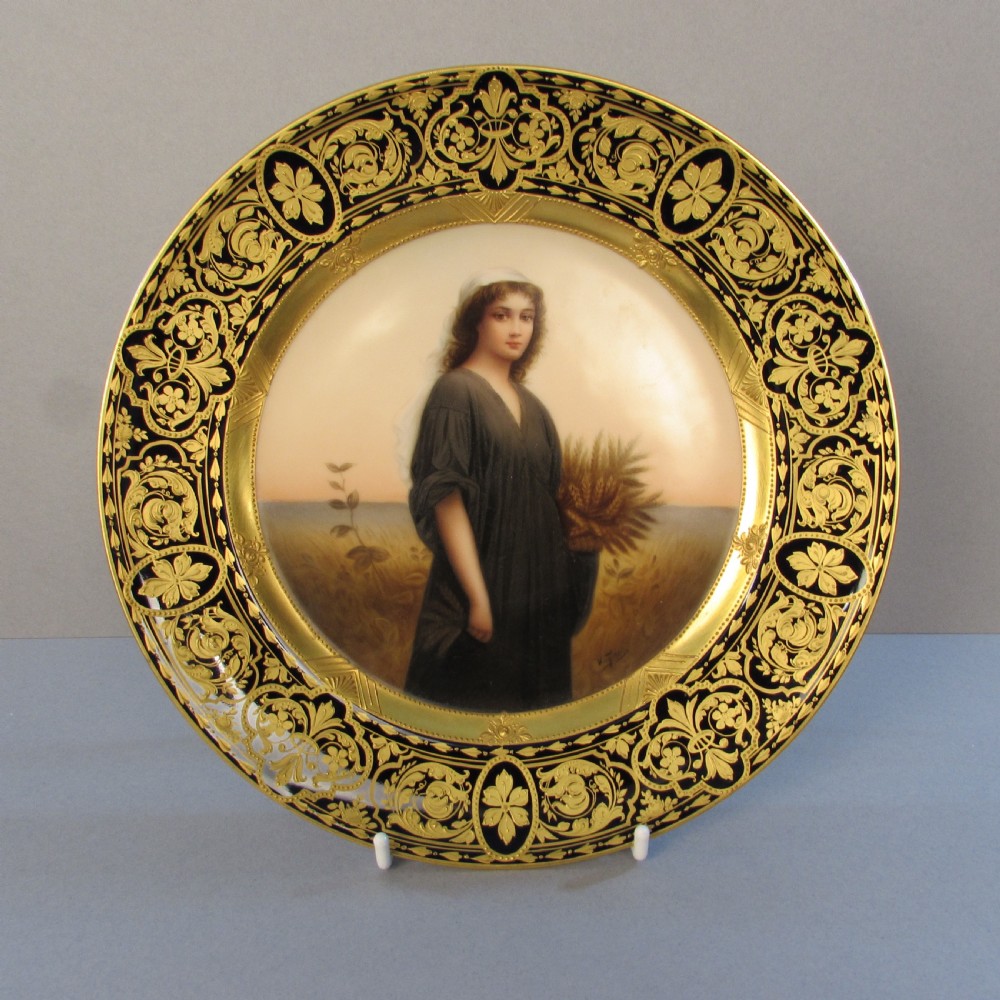 A circular porcelain cabinet plate featuring a central hand-painted portrait of a woman holding wheat, framed by a broad black rim decorated with gold leaf scrollwork.
