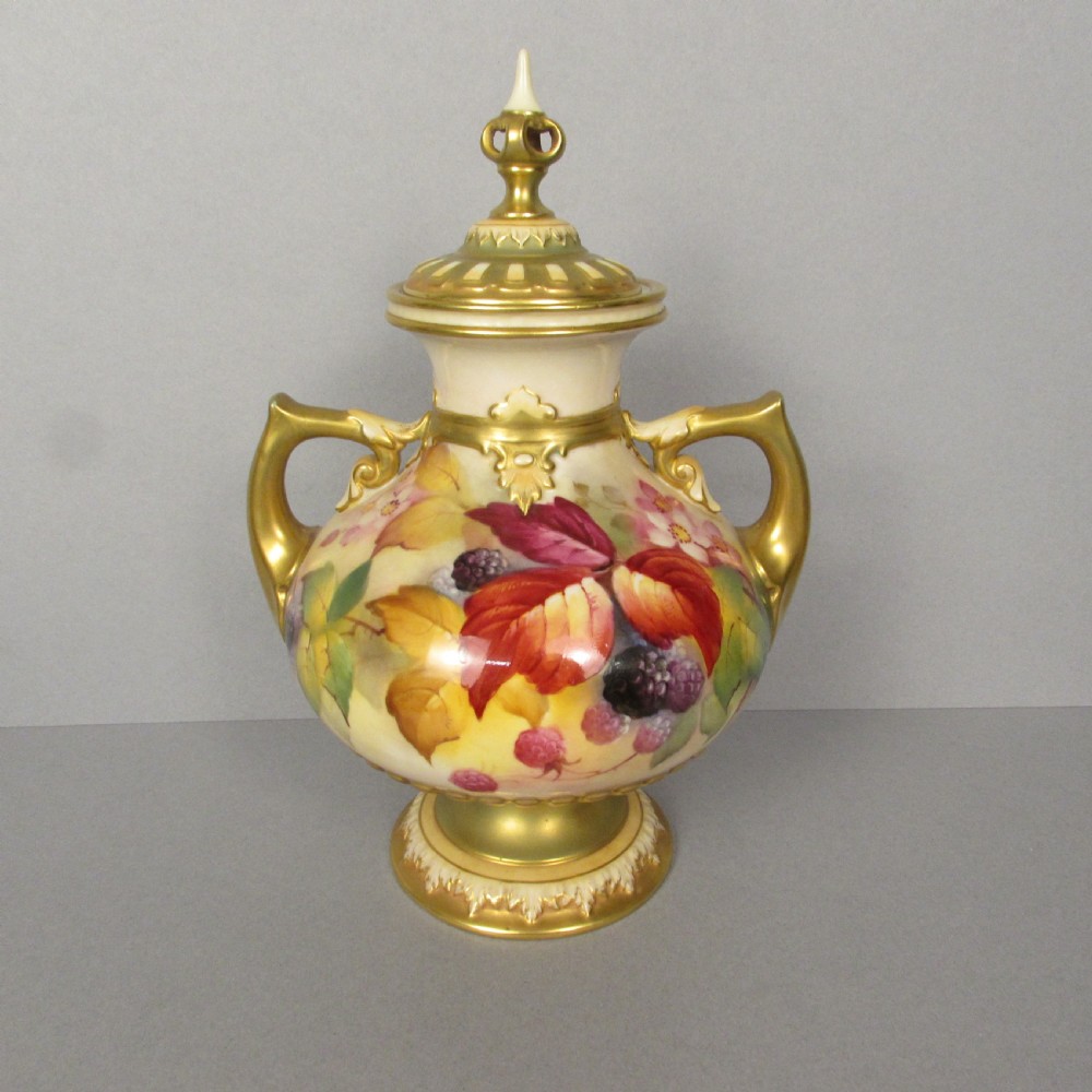 A twin-handled porcelain urn with a finial lid, featuring hand-painted autumn foliage and fruit decoration with rich gilding on the handles, neck, and base.