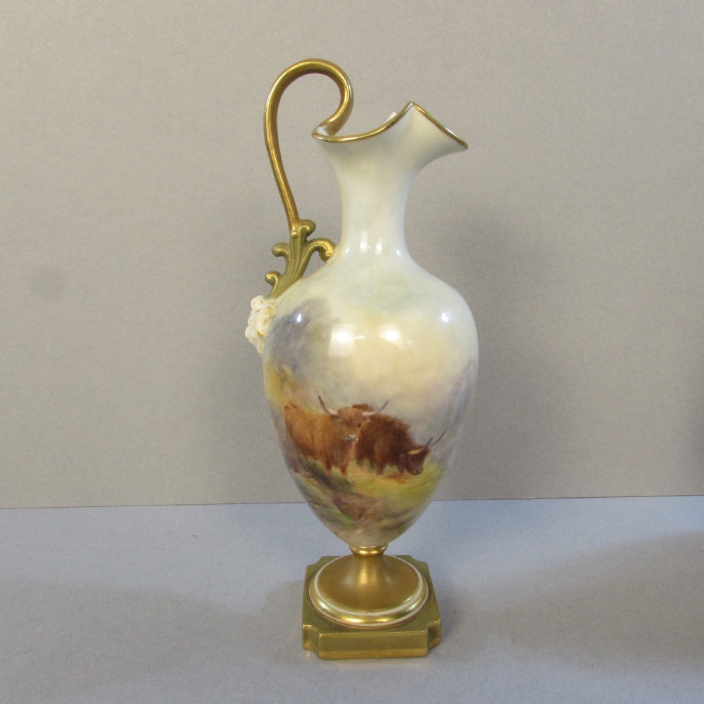An ornate porcelain ewer featuring a hand-painted scene of Highland cattle, a flared spout, a gilded scroll handle, and a square gold-finished base.