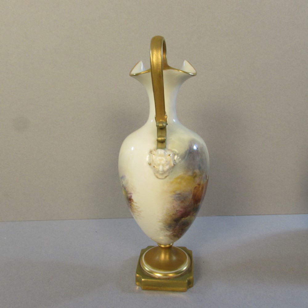 An ornate porcelain ewer featuring a hand-painted scene of Highland cattle, a flared spout, a gilded scroll handle, and a square gold-finished base. View II