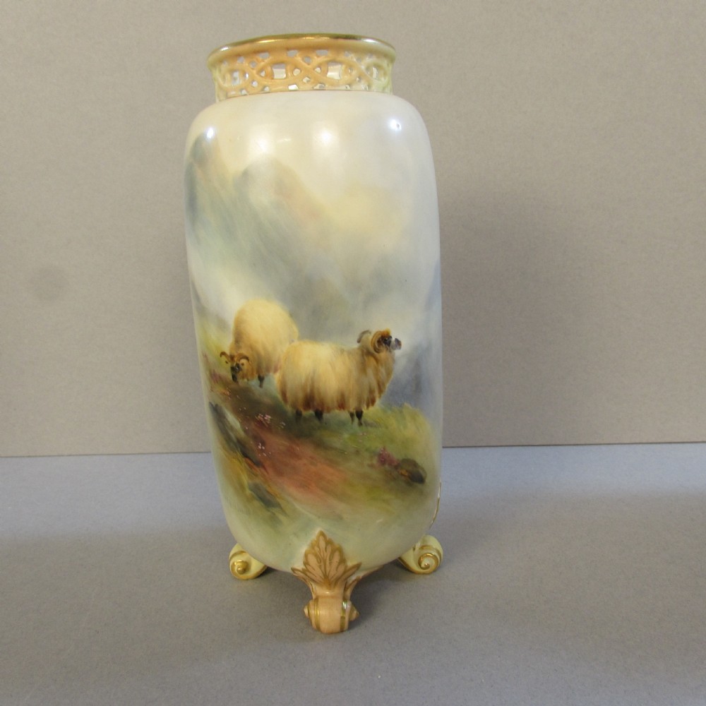 A cylindrical soft cream porcelain vase decorated with a painted scene of Highland sheep, featuring a reticulated gilt rim and four gilded scroll feet.