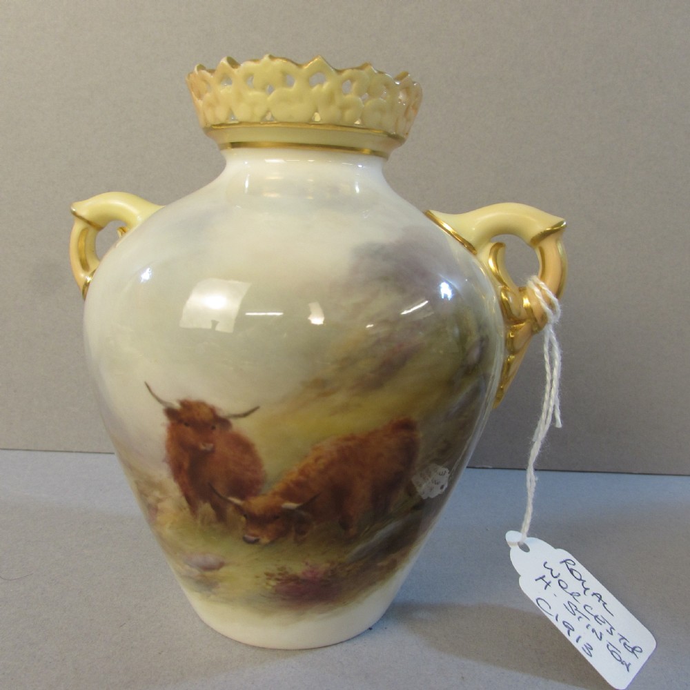 A cream porcelain vase with reticulated gilt rim, twin handles, and a hand-painted scene depicting Highland cattle in a misty landscape.