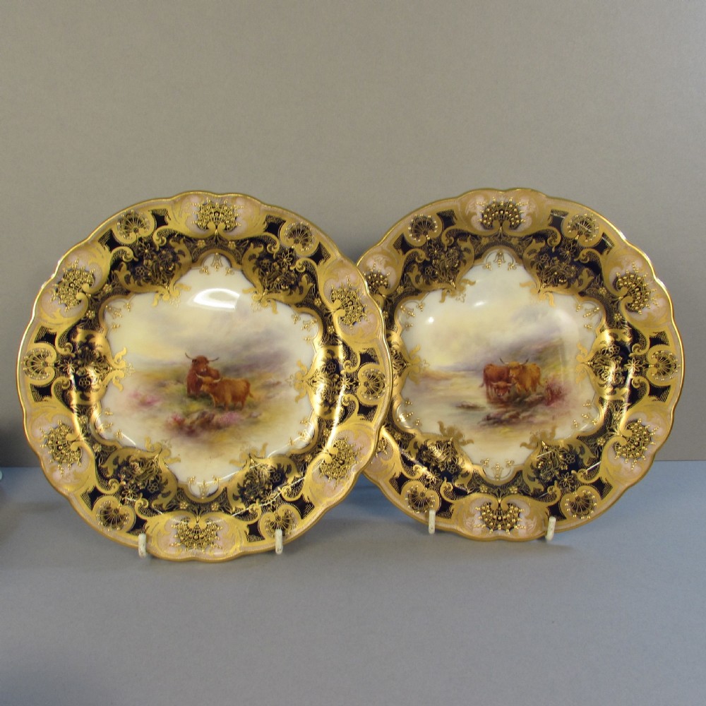 A pair of round antique Royal Worcester porcelain cabinet plates featuring hand-painted Highland cattle landscape scenes encircled by elaborate gilded cobalt blue borders.