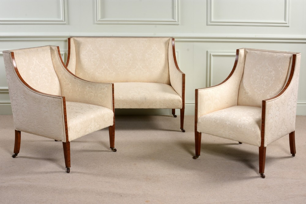 An antique three-piece Edwardian seating suite comprising a two-seater sofa and two matching armchairs upholstered in cream damask with mahogany frames and brass castors.